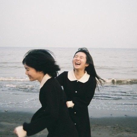 two women walking on the beach with one laughing and another looking up at the sky