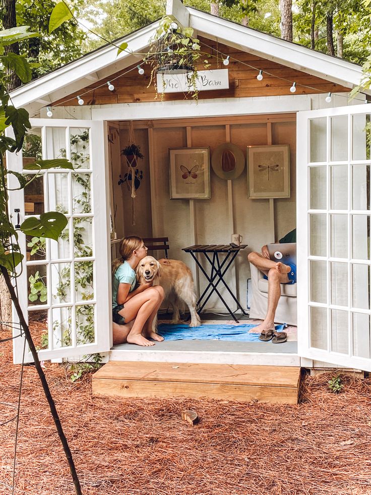 two people and a dog are sitting in a small white shed with windows on the side