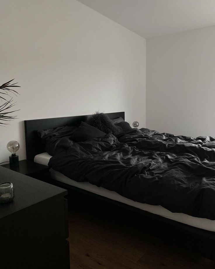 an unmade bed with black sheets and pillows in a white room next to a window