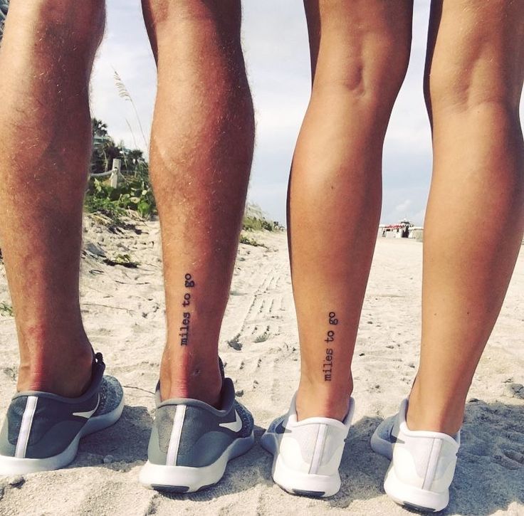 two people standing on the beach with their feet in the sand and one has tattoos on his left leg