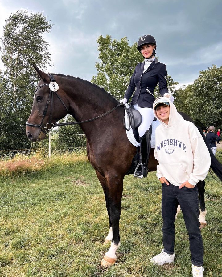 two people standing next to each other on top of a brown horse in a field