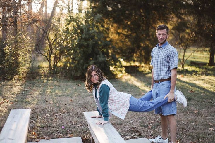 a man and woman standing on top of a wooden bench in the grass with trees behind them