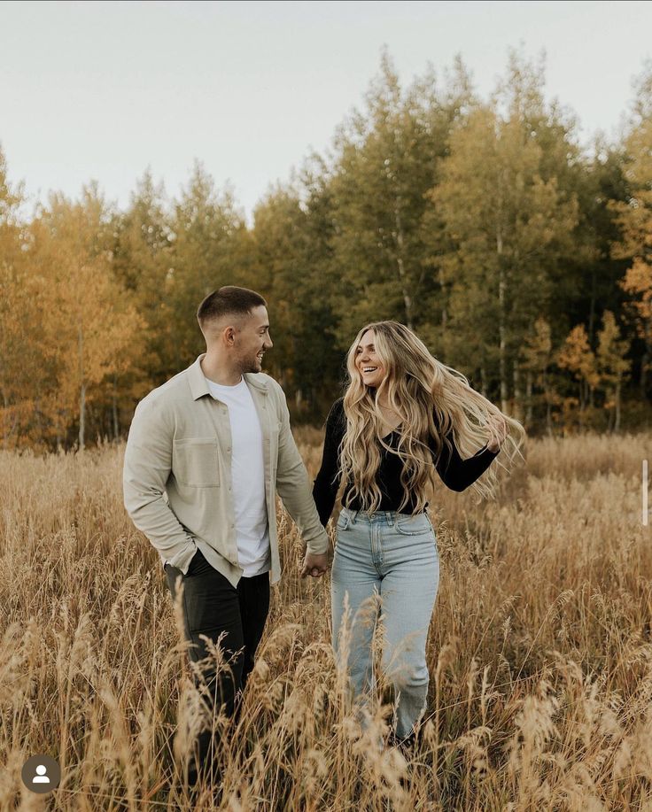 a man and woman holding hands walking through tall grass with trees in the back ground