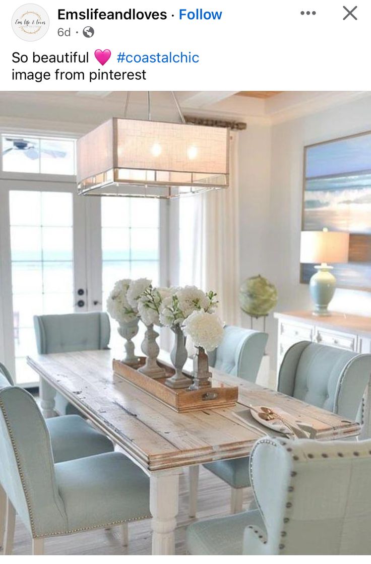 a dining room table with blue chairs and white flowers on the centerpiece, in front of a large window