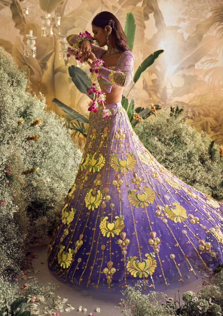 Step into a world of enchantment with our shaded lilac tulle lehenga set - a spellbinding fusion of colors and textures. Watch as custom cut acrylic pieces, gold metal embellishments, and rich fabric appliques come together to create a truly mesmerizing design. Paired with our signature butterfly full-sleeved blouse in lilac silks, and a delicately embroidered tulle dupatta that glistens with metals, pearls, and sequins. Get ready to dazzle and make a statement on your special day! Designer Purple Organza Sharara, Purple Sets With Sheer Dupatta For Reception, Lavender Embroidered Sharara For Wedding, Purple Reception Sets With Sheer Dupatta, Lavender Sharara With Intricate Embroidery For Wedding, Reception Sets With Purple Sheer Dupatta, Traditional Hand Embellished Purple Dress, Festive Purple Hand Embellished Sets, Festive Hand Embellished Purple Sets