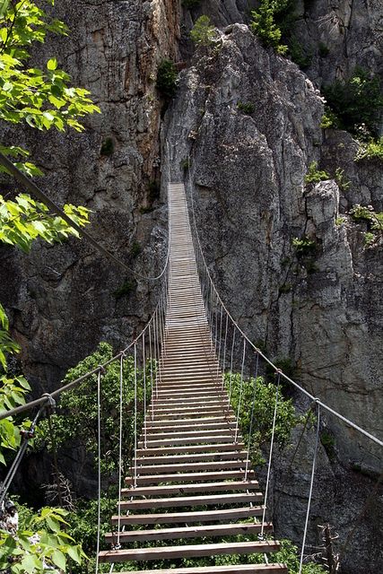 a wooden suspension bridge over a river next to a rocky mountain side with trees on both sides