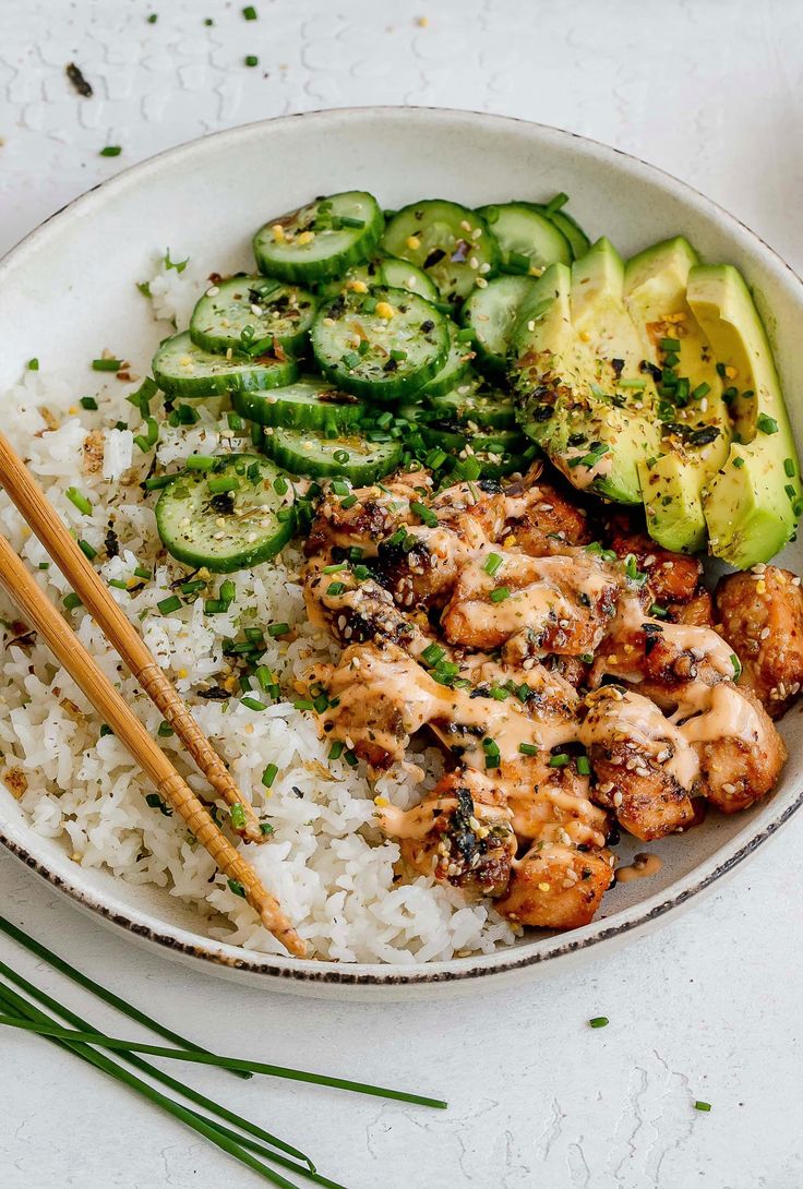 a white bowl filled with rice and chicken next to cucumber, avocado and chopsticks