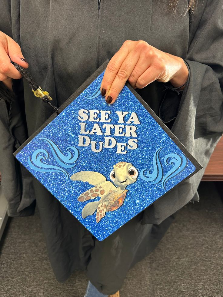 a person holding a blue graduation cap with a sea ya later dude design on it