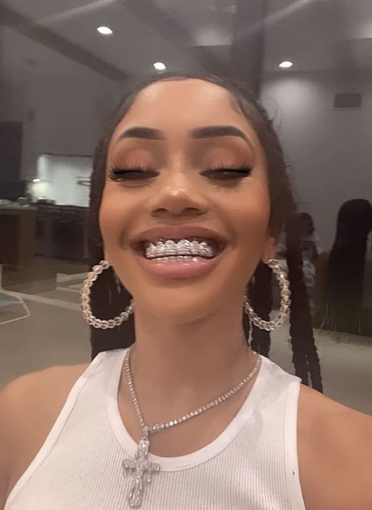 a smiling woman with braces on her teeth in an office setting, wearing a white tank top and large hoop earrings