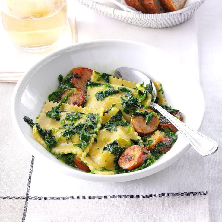 a white bowl filled with pasta and spinach on top of a table next to bread