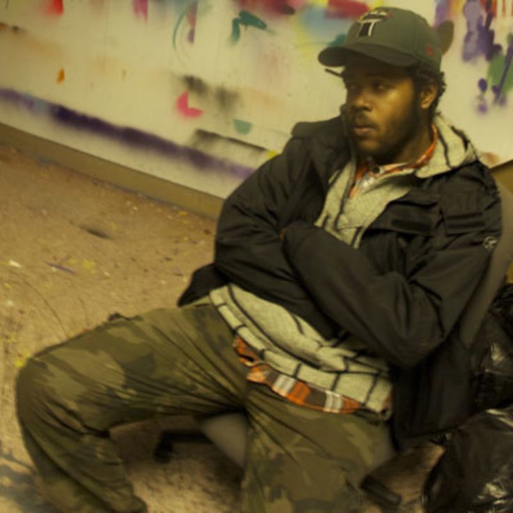 a man sitting on the ground in front of a wall with graffiti and trash bags