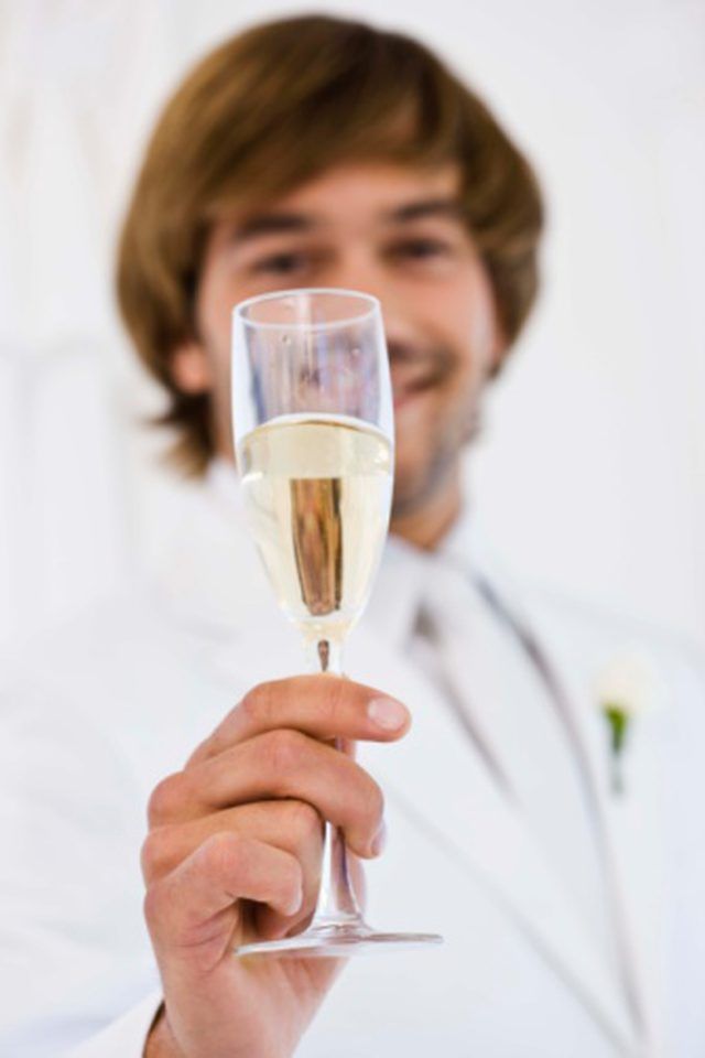 a man in a white suit holding a glass of wine and smiling at the camera