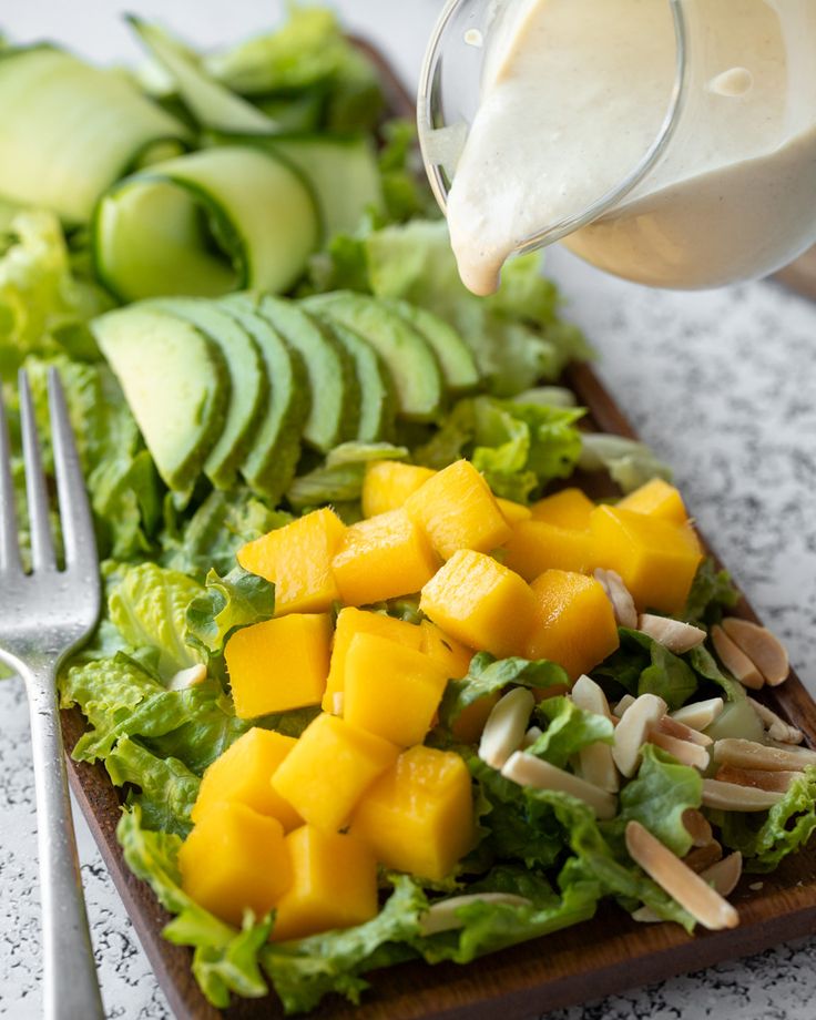 a person pouring dressing onto a salad with avocado and mangoes on the side