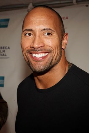 a bald man smiling at the camera in front of a white wall with a sign behind him