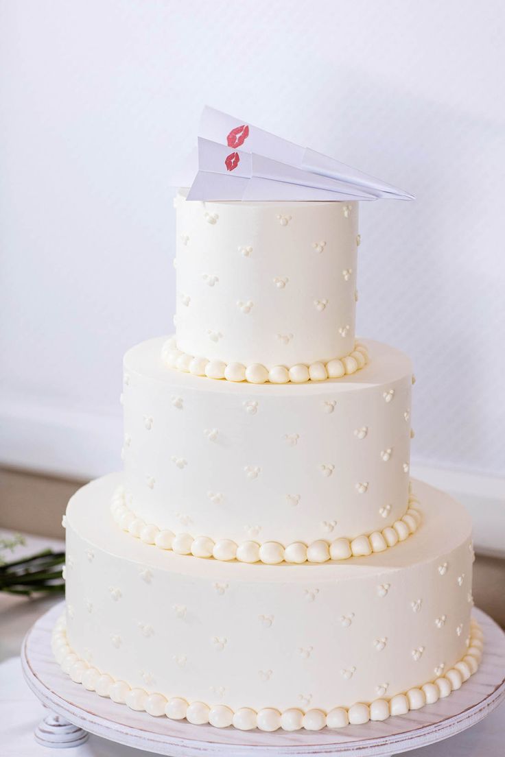 a three tiered cake with white frosting and pearls on the bottom is topped with a paper airplane