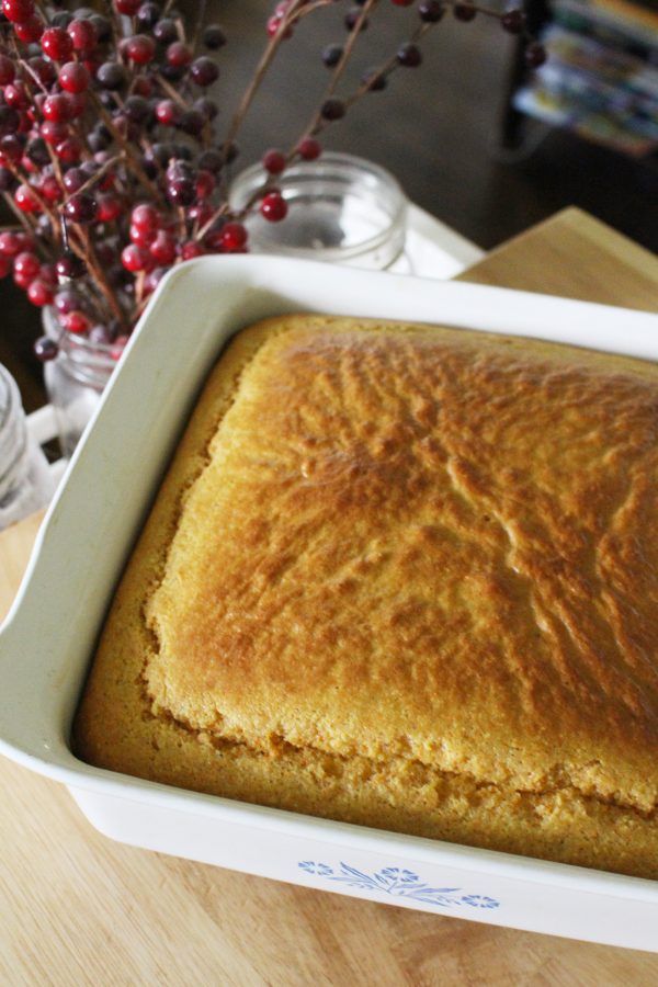 a square cake in a white dish on a wooden table next to a vase with red berries