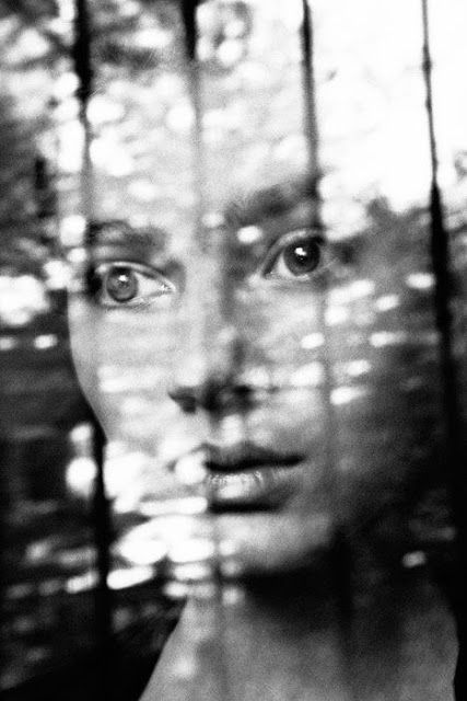 a black and white photo of a woman's face through a window with trees in the background