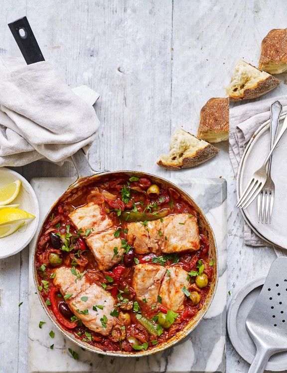 a pan filled with fish and vegetables on top of a wooden table next to bread