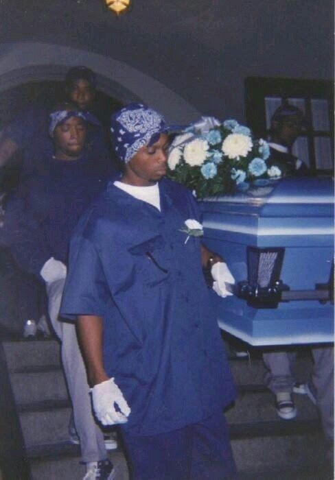 a man standing next to a blue casket with flowers in it and other people around him