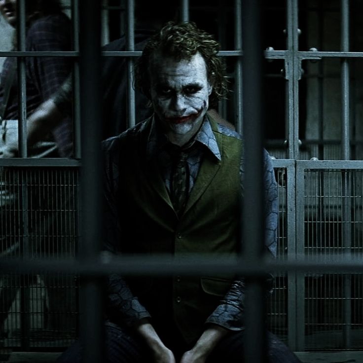 the joker is sitting behind bars in his caged area, looking out at another person