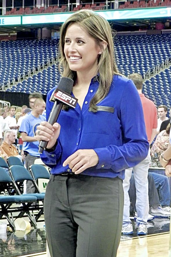 a woman in blue shirt and black pants holding a microphone while standing on a basketball court