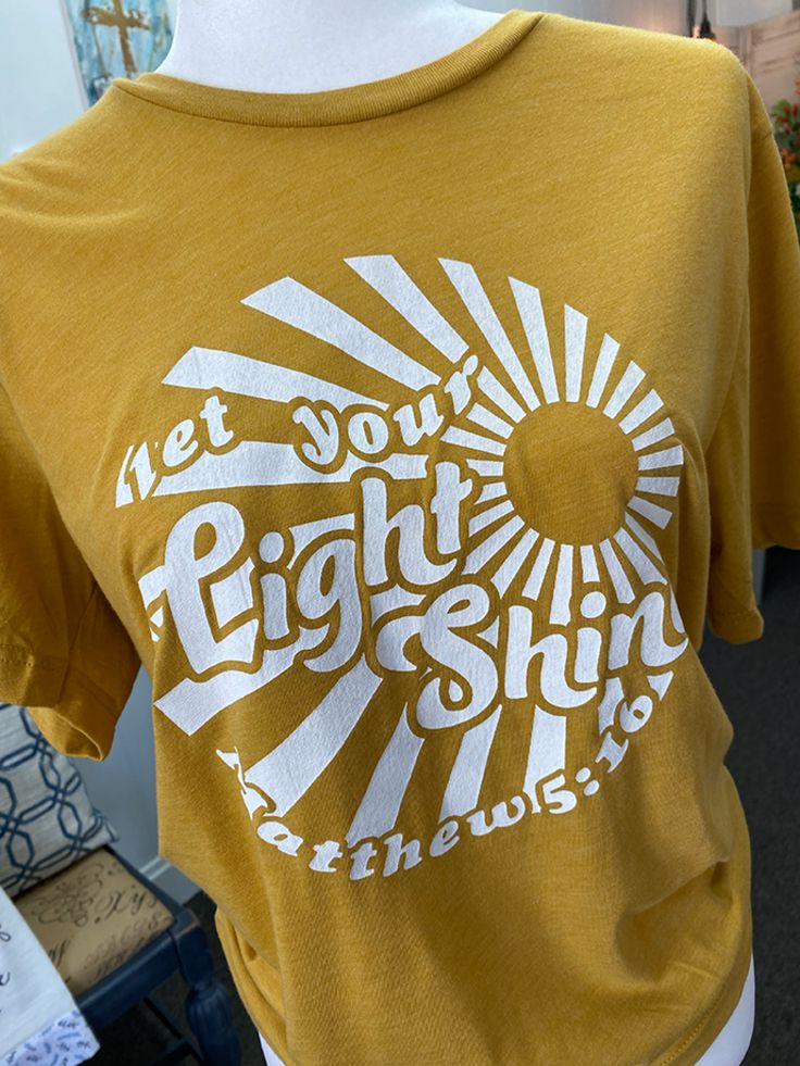 Seasons Inspirations’ Christian t-shirts will enable you to share the gospel with hundreds of people. Wear your faith proudly while being comfortable & stylish in one of the many Christian tees that we have to offer. Scripture reference: John 16:33 “In the same way, let your light shine before others, that they may see your good deeds and glorify your Father in heaven.” Our Let Your Light Shine t shirts are screen-printed by hand on a Bella & Canvas short sleeve Mustard shirt. Available in sizes Fca Shirt Ideas, Let Your Light Shine Shirt, Sunshine Shirt Ideas, Be The Light Craft, Shine Theme, Youth Group Shirts, Mission Trip Shirts, Stellar Vbs, Christian Girlie