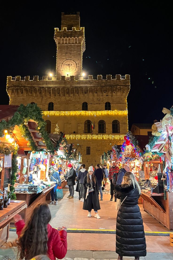 people are walking around an outdoor market with lights on the buildings and clock tower in the background