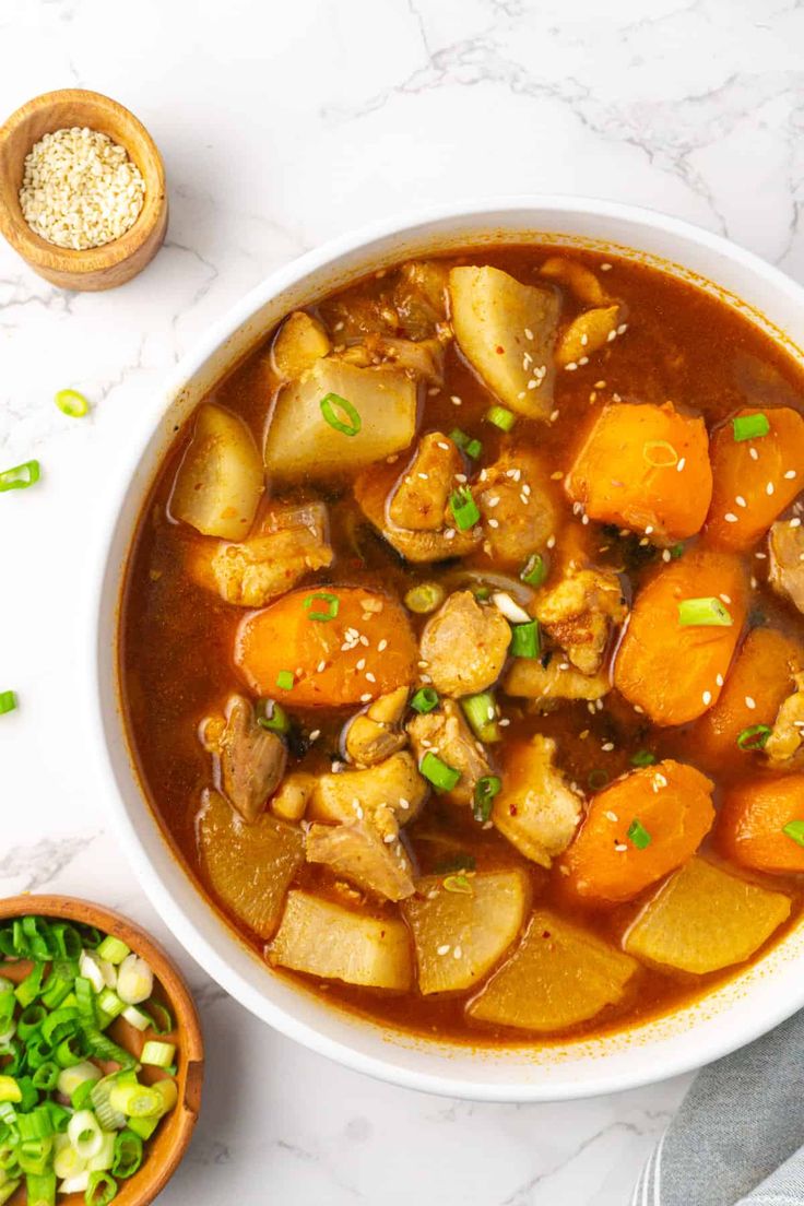 a bowl of stew with carrots, potatoes and green onions on the side next to a wooden spoon