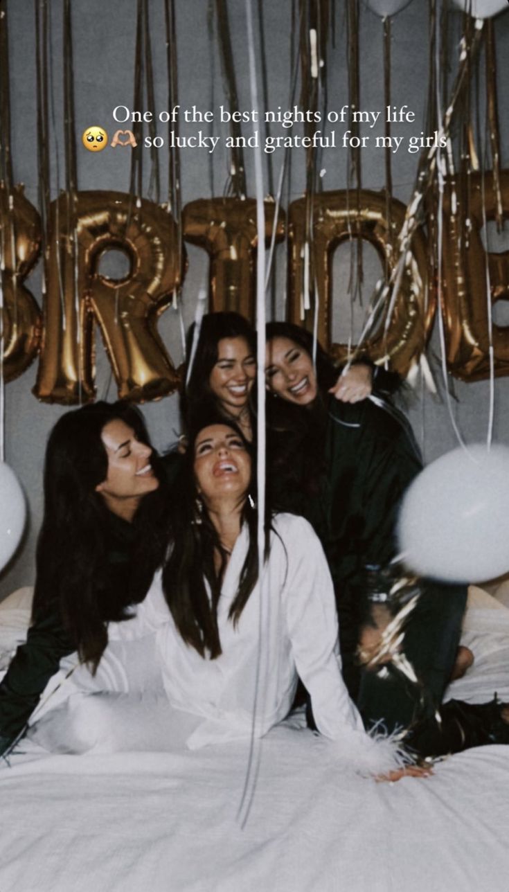 four women sitting on a bed with balloons and streamers in the air behind them