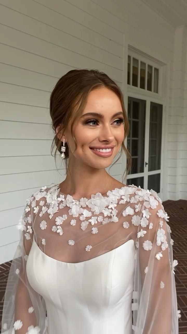 a woman wearing a white dress with flowers on the shoulders and sleeves, standing in front of a house