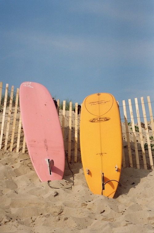 two surfboards sitting on the beach next to a white picket fence and wooden posts