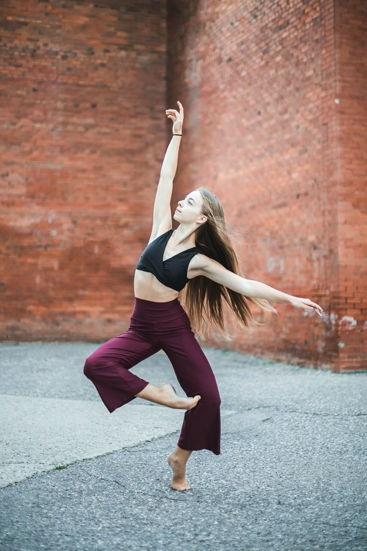 a woman in black top and purple pants doing a dance pose on the street with brick wall behind her