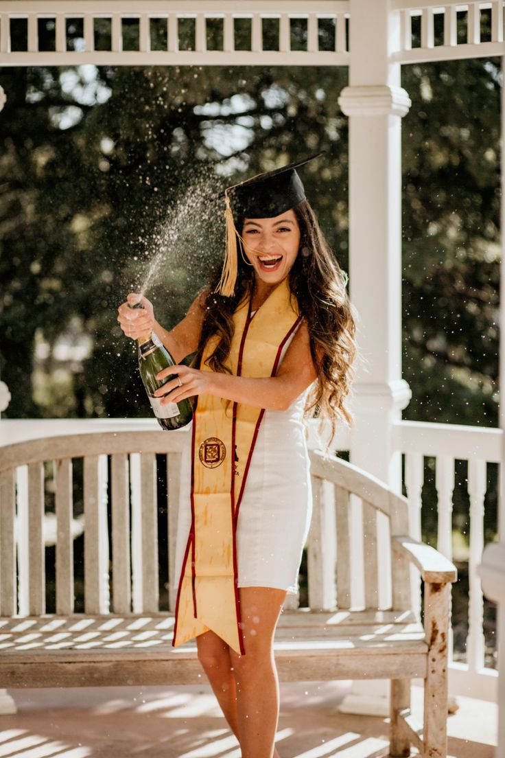 a woman in graduation gown holding a bottle and throwing it into the air while standing on a porch