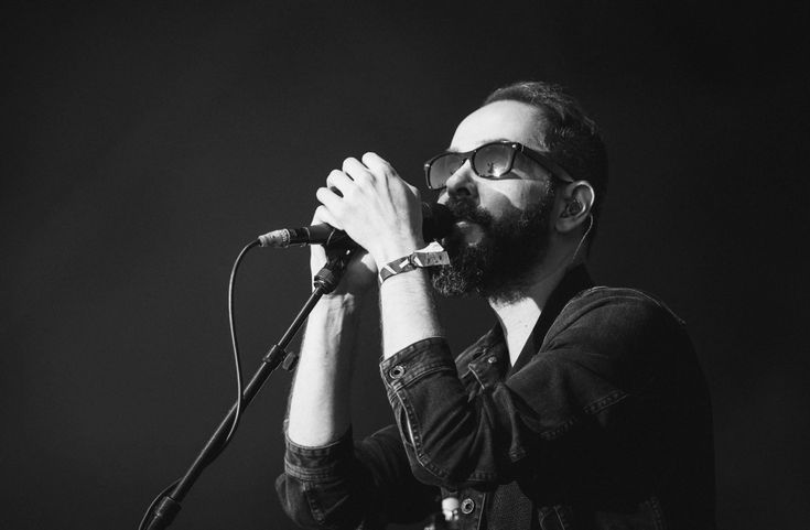 a bearded man with glasses on singing into a microphone in front of a black background