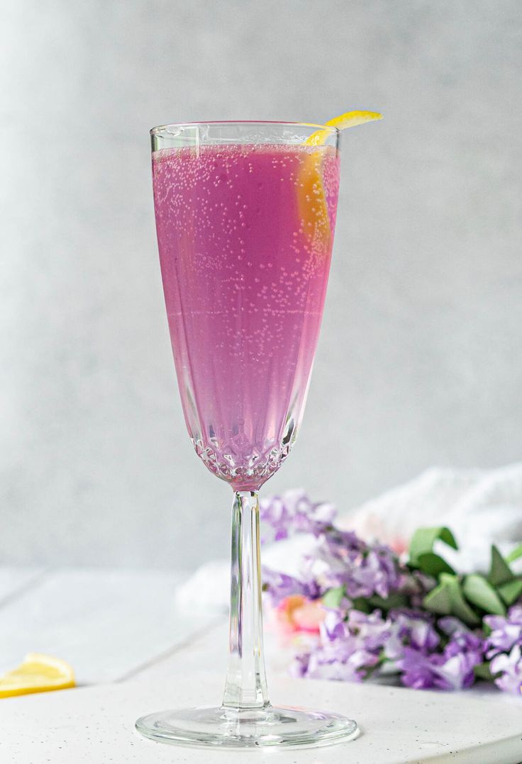 a glass filled with pink liquid next to purple flowers and lemon wedges on a white surface