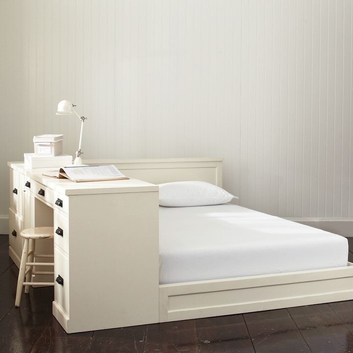 a small white bed sitting next to a desk with a lamp on top of it