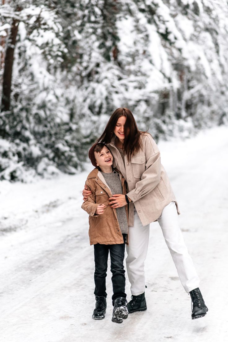 a mother and son walking in the snow on a path with pine trees behind them