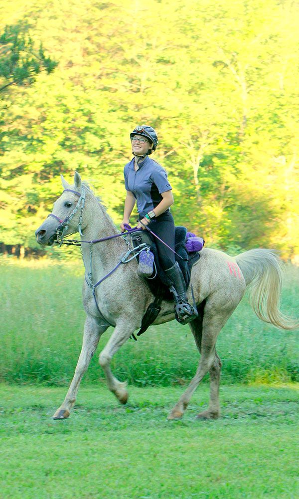 a woman riding on the back of a white horse through a lush green field with trees in the background