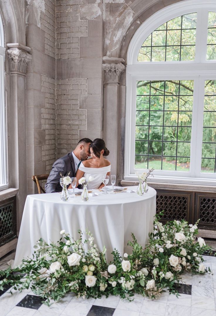 a bride and groom sitting at a round table with greenery in front of them