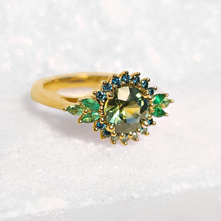 Make a statement with this one-of-a-kind stunner!  It would be ideal as an alternative engagement ring or an everyday cocktail ring.  It is made of eco-friendly 14-karat yellow gold and set with a magnificent Montana sapphire.  The sapphire provides hues of green and yellow and It is accented with blue diamonds and emeralds.  If you are a lover of greenish-blue this 14K Montana Sapphire, Blue Diamond, and Emerald Ring is a match for you! Green Emerald Cut Sapphire Ring, Green Sapphire Rings With Prong Setting, Green Sapphire Ring With Center Stone, Green Diamond Ring With Center Stone For Proposal, Green Sapphire Ring With Halo Setting For Promise, Green Sapphire Round Ring, Fine Jewelry Green Sapphire Ring With Prong Setting, Green Sapphire Ring With Accent Stones For Promise, Green Sapphire Promise Ring With Accent Stones