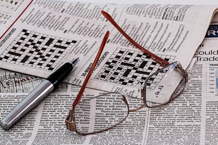 a newspaper with glasses, pen and eyeglasses laying on it next to a crossword puzzle