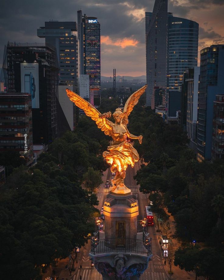 the angel statue is lit up at night in front of tall buildings and skyscrapers