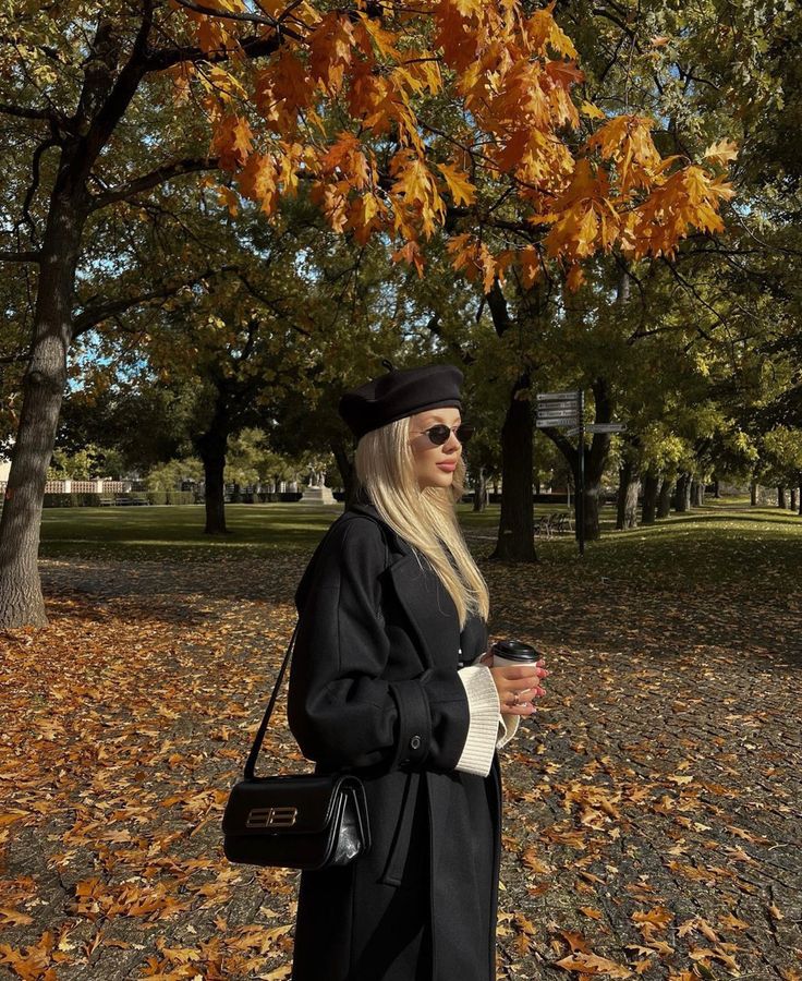 a woman standing under a tree with leaves on the ground and holding a coffee cup