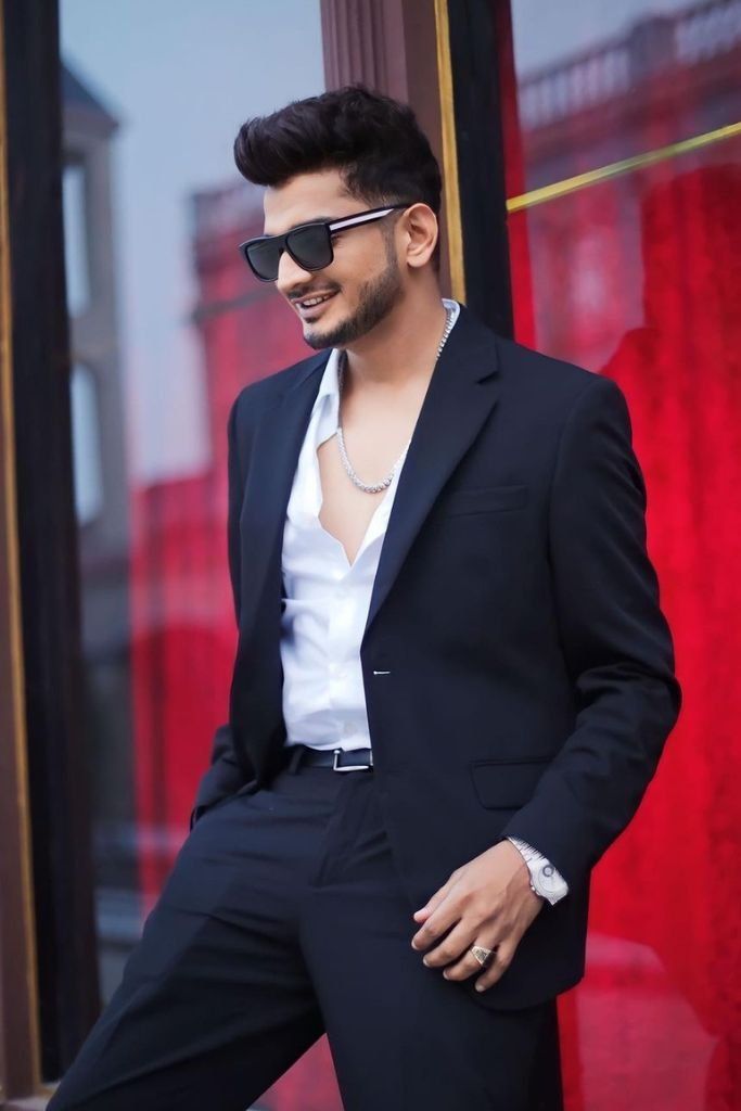 a man in a black suit and sunglasses leaning against a red wall with his hand on his hip