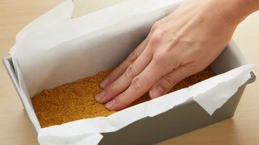 a hand reaching into a box filled with sand on top of a wooden table next to a paper bag