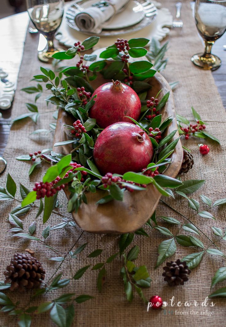 two apples are sitting in a bowl on a table with greenery and pine cones