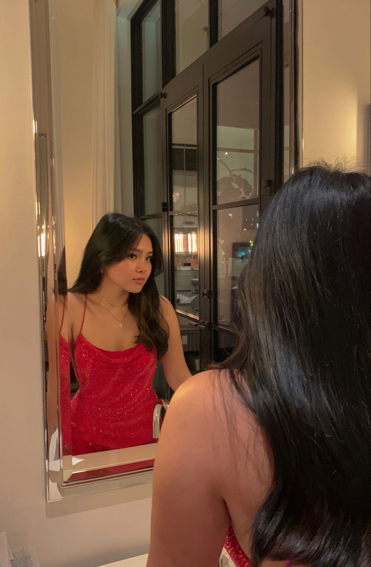 a woman in a red dress looking at herself in the mirror with long black hair