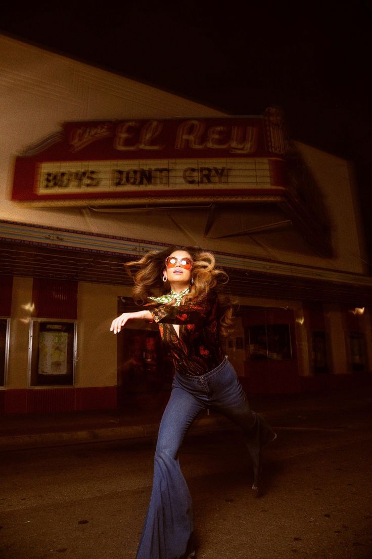 a woman is dancing in front of a movie theater with her hair blowing in the wind