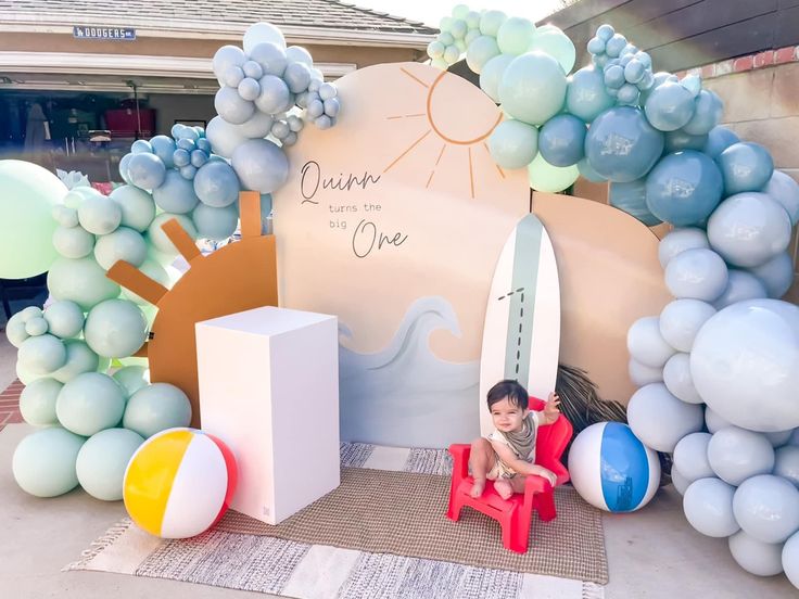 a little boy sitting on a red chair in front of a surfboard and balloon arch