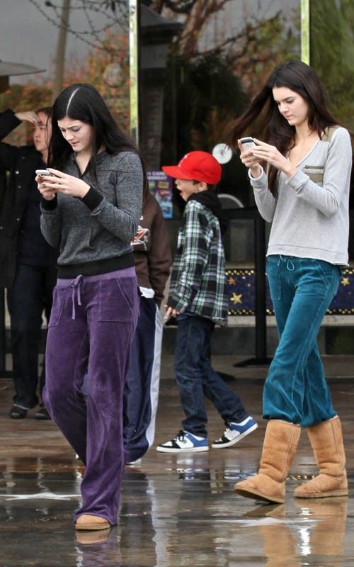 two women standing in the rain looking at their cell phones while others look on with umbrellas